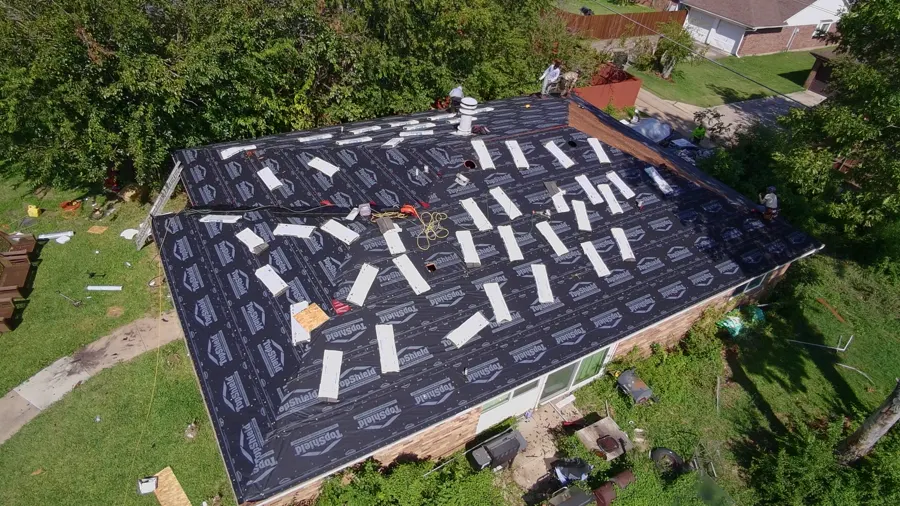 Aerial view of a single-story brick home under black synthetic underlayment, shingle bundles staged across the deck